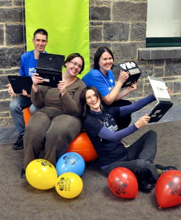 Four volunteers sitting together holding laptops, with Software Freedom Day balloons around them.